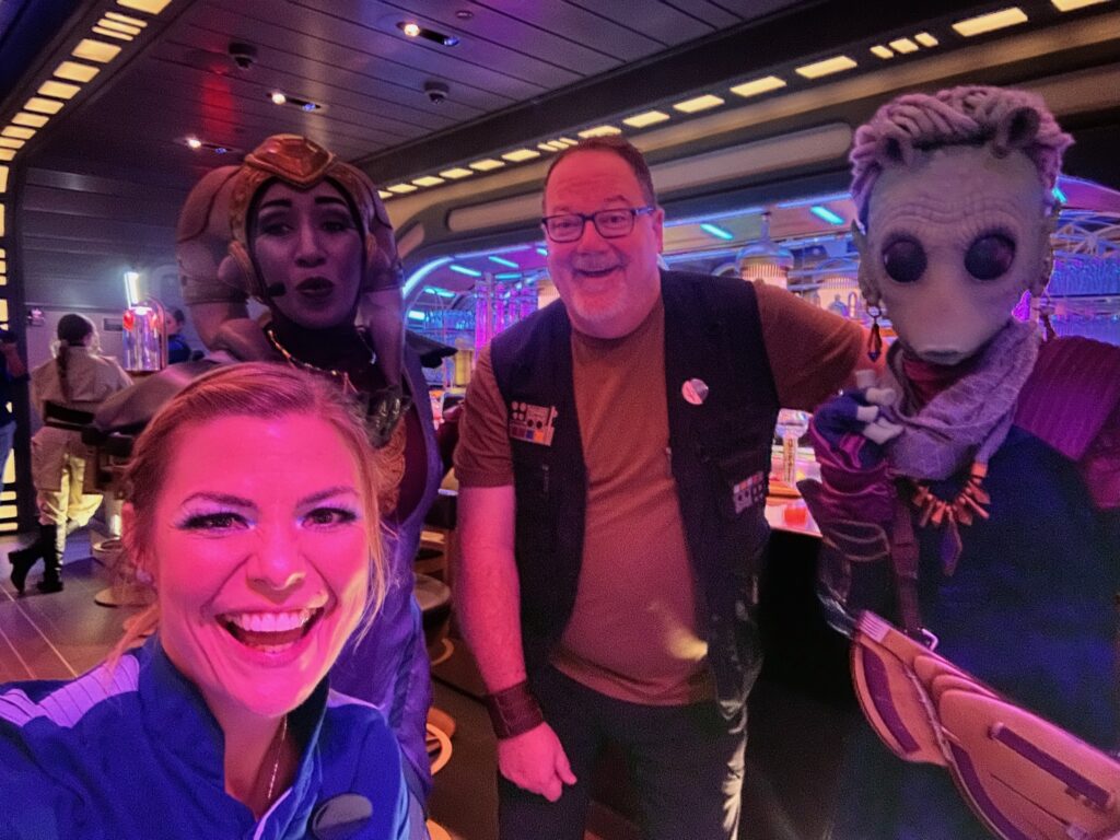 Jeff in star wars outfit in the Sublight Lounge on the Star Wars:Galactic Starcruiser, posed with Gaya, Oouanni, and a crew member
